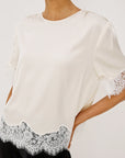 An off-white short-sleeve blouse with delicate white lace trim at the hem and sleeve cuffs, worn by a model against a light gray studio background.