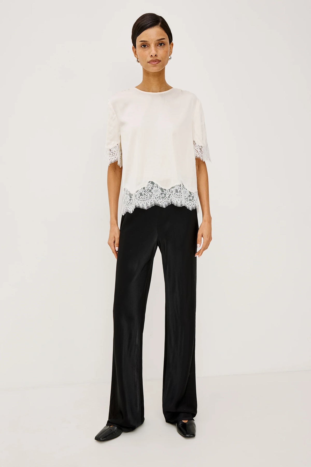 A model stands against a plain white studio background wearing a white short-sleeve top with black lace trim at the hem and black wide-leg trousers.