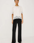 A model stands against a plain white studio background wearing a white short-sleeve top with black lace trim at the hem and black wide-leg trousers.