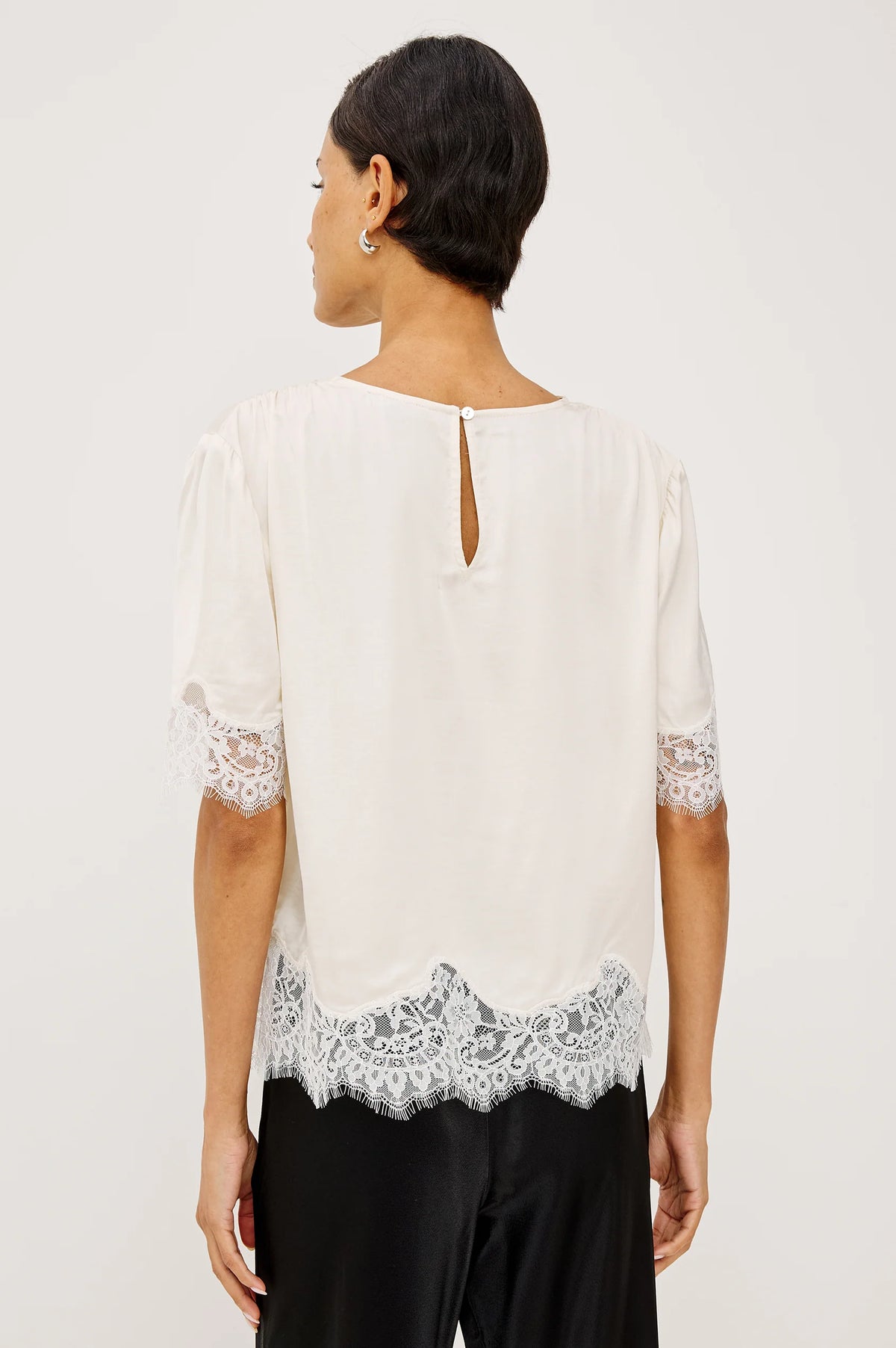 A woman shown from the back wearing a white blouse with short puff sleeves, lace trim at the cuffs and hem, and a small keyhole cutout at the neckline, paired with black pants against a light gray studio background.