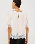A woman shown from the back wearing a white blouse with short puff sleeves, lace trim at the cuffs and hem, and a small keyhole cutout at the neckline, paired with black pants against a light gray studio background.