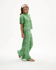 A woman modeling a sage green two-piece linen set with embroidered leaf patterns, featuring a short-sleeve top and matching wide-leg pants, against a plain white studio background.