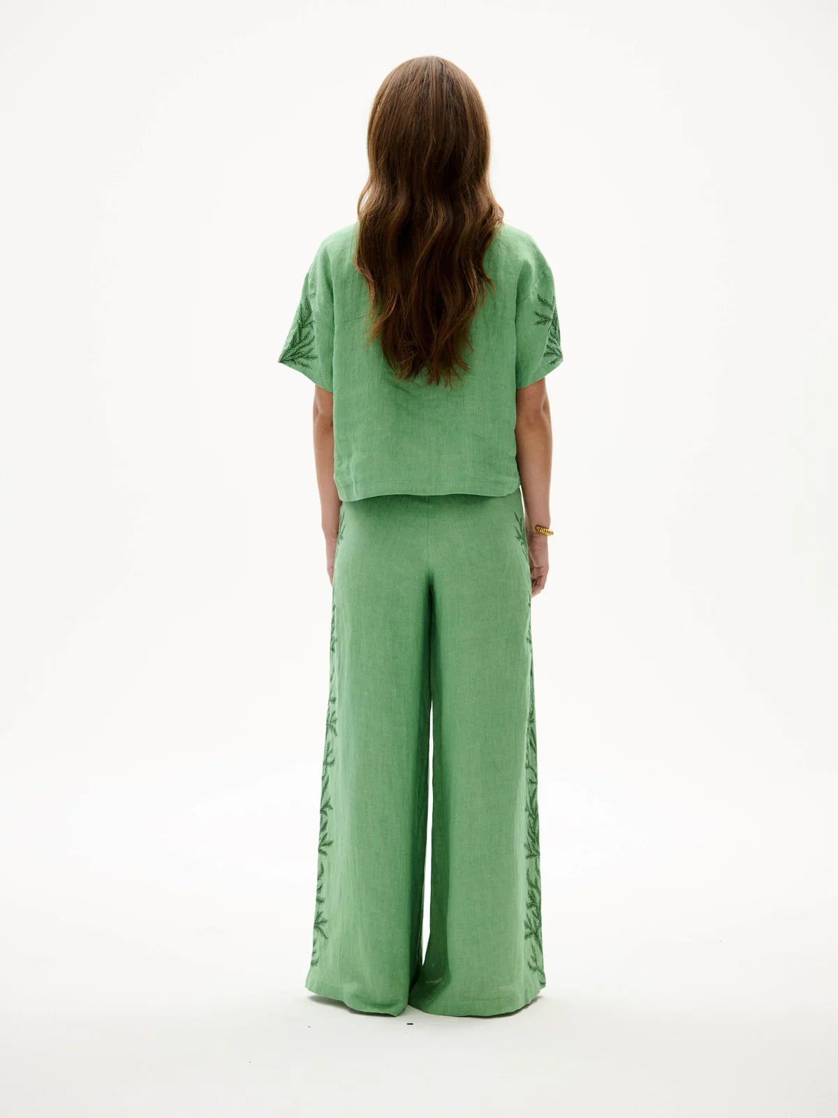 A woman viewed from behind wearing a sage green two-piece linen set with a short-sleeve cropped top and wide-leg trousers, featuring decorative lace-up detailing along the sides of the pants.