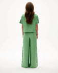 A woman viewed from behind wearing a sage green two-piece linen set with a short-sleeve cropped top and wide-leg trousers, featuring decorative lace-up detailing along the sides of the pants.