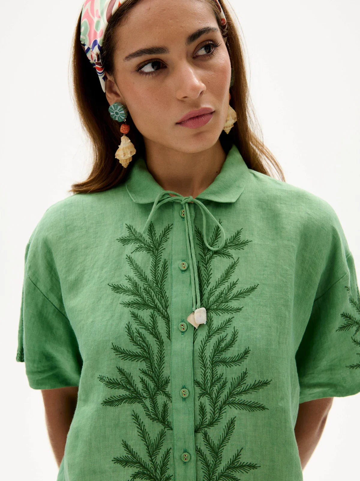 A woman wearing a green short-sleeve collared shirt with dark green embroidered leaf patterns down the front and a heart-shaped button, photographed against a plain light background.