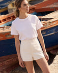 Woman in a white top and beige shorts standing among old boats.