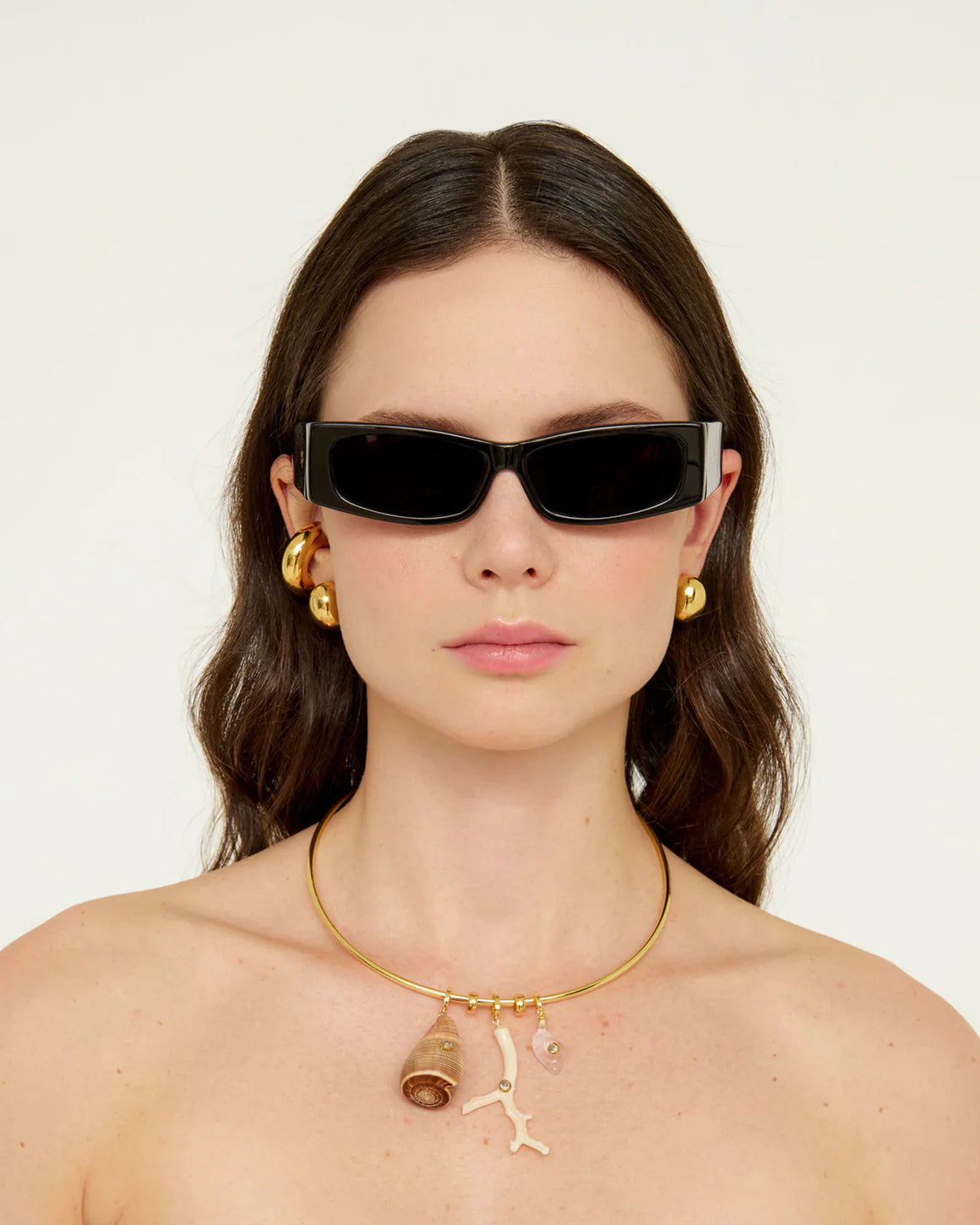 Woman wearing black sunglasses and gold jewelry against a white background