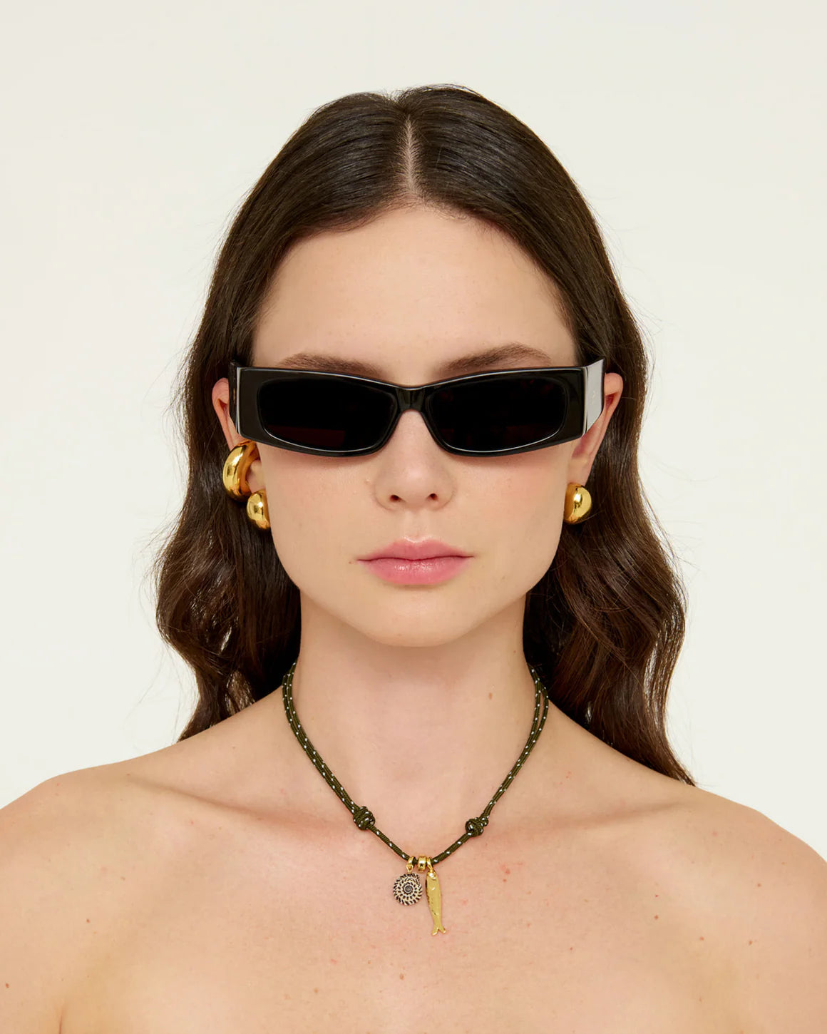 Woman wearing black sunglasses and gold earrings  and green cord charm necklace with a plain background