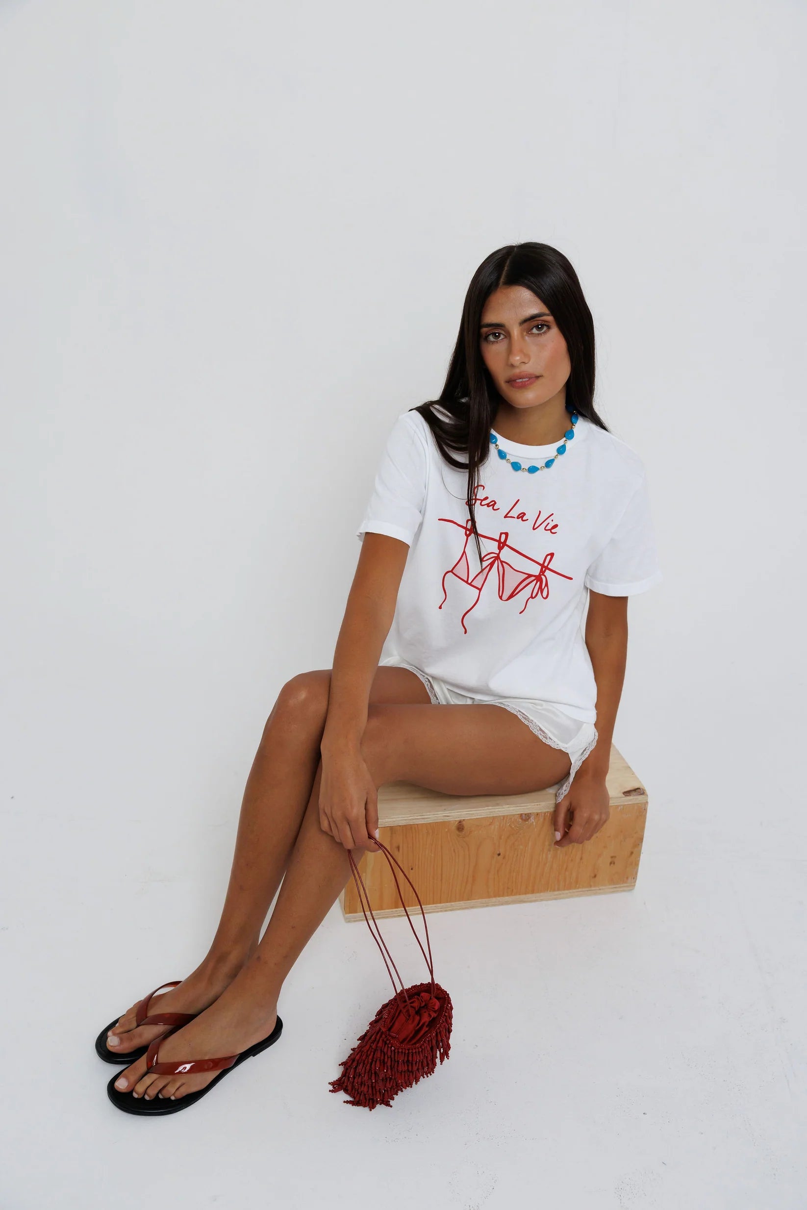 model sitting while wearing sea la ie graphic tee with a turquoise necklace on a white background