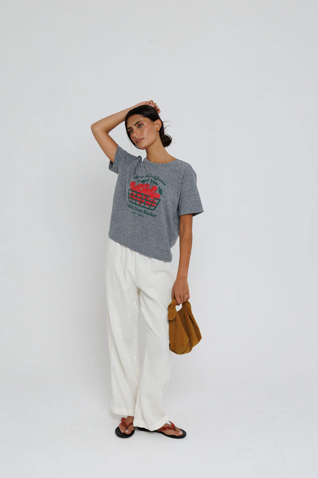 Woman wearing a gray t-shirt with red text and white pants, holding a brown bag on a white background