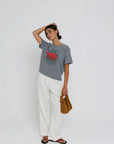 Woman wearing a gray t-shirt with red text and white pants, holding a brown bag on a white background