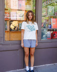 Woman wearing a white t-shirt with text and blue shorts standing in front of a store window.