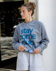 Woman wearing a gray sweatshirt with 'stay home club' text, holding sunglasses, standing against a building.