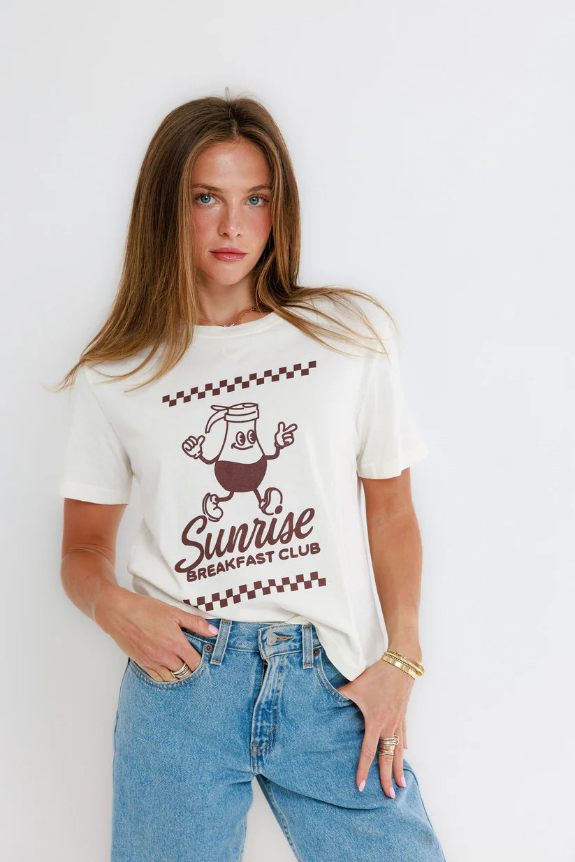 Woman wearing a white t-shirt with 'Sunrise Breakfast Club' design and blue jeans on a white background