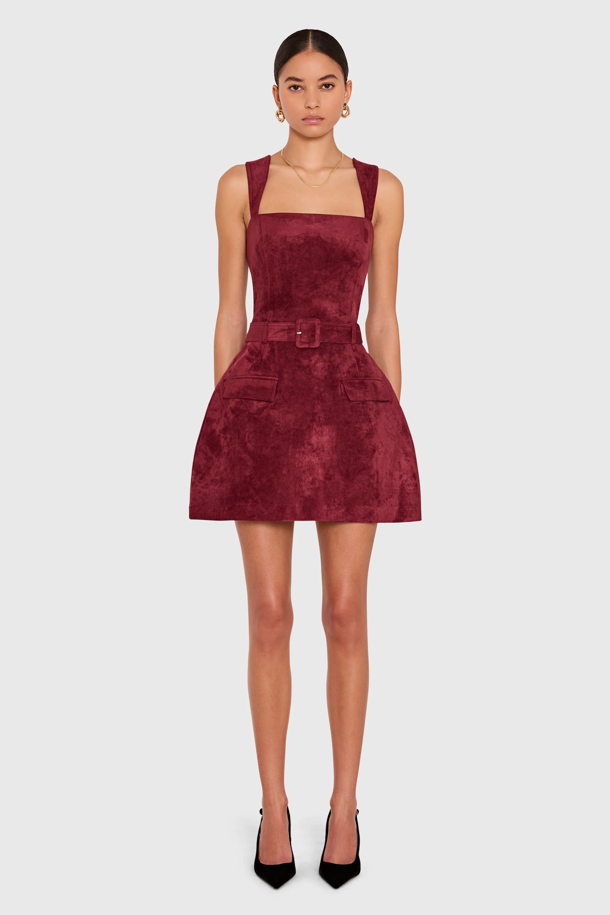 A model wearing a sleeveless burgundy velvet mini dress with a square neckline, wide shoulder straps, a matching belt with a square buckle, and front patch pockets, paired with black high heels against a light gray background.