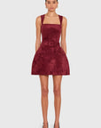 A model wearing a sleeveless burgundy velvet mini dress with a square neckline, wide shoulder straps, a matching belt with a square buckle, and front patch pockets, paired with black high heels against a light gray background.