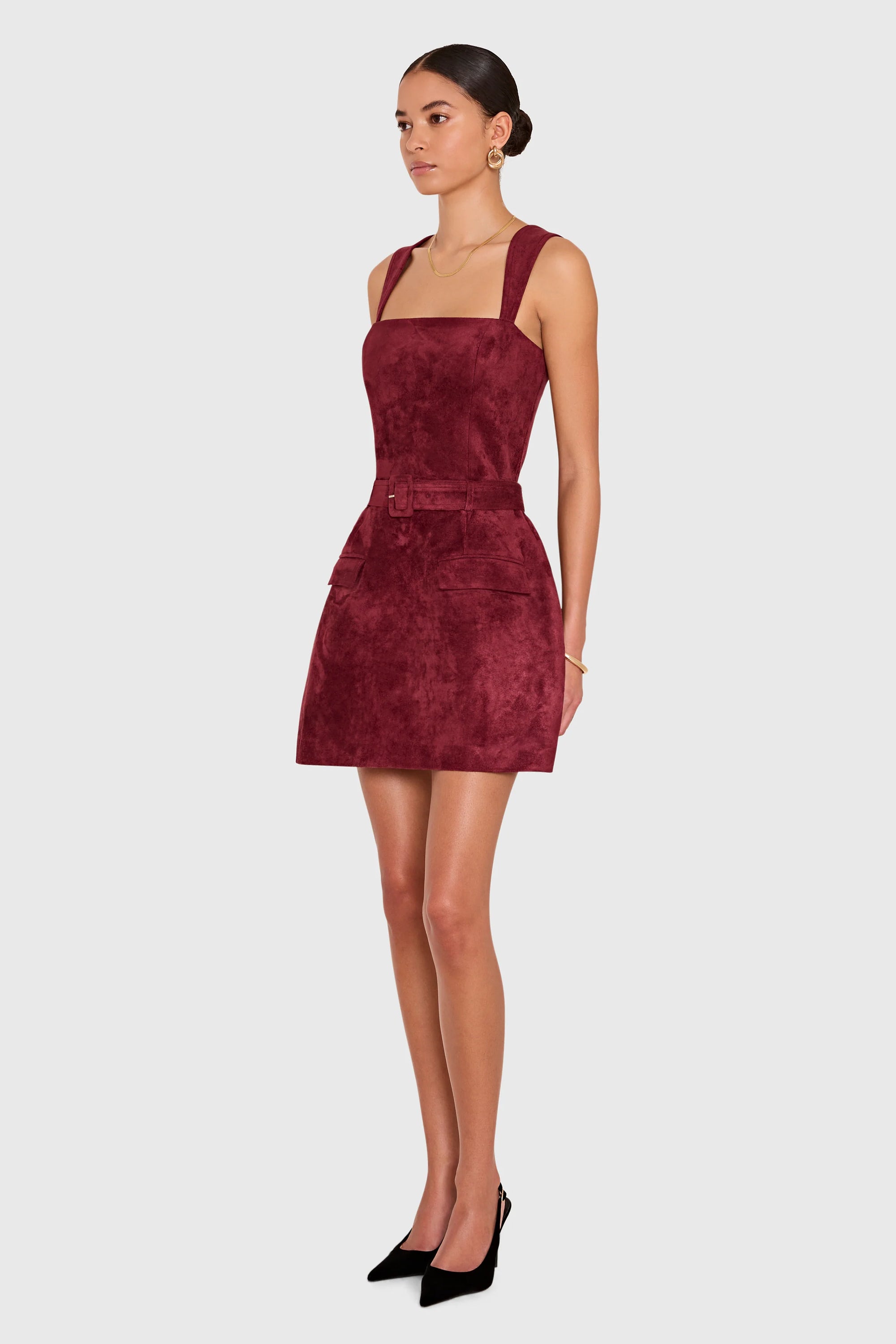 A woman modeling a sleeveless, square-neck mini dress in a deep burgundy velvet fabric, styled with black pointed-toe heels against a solid light gray background.