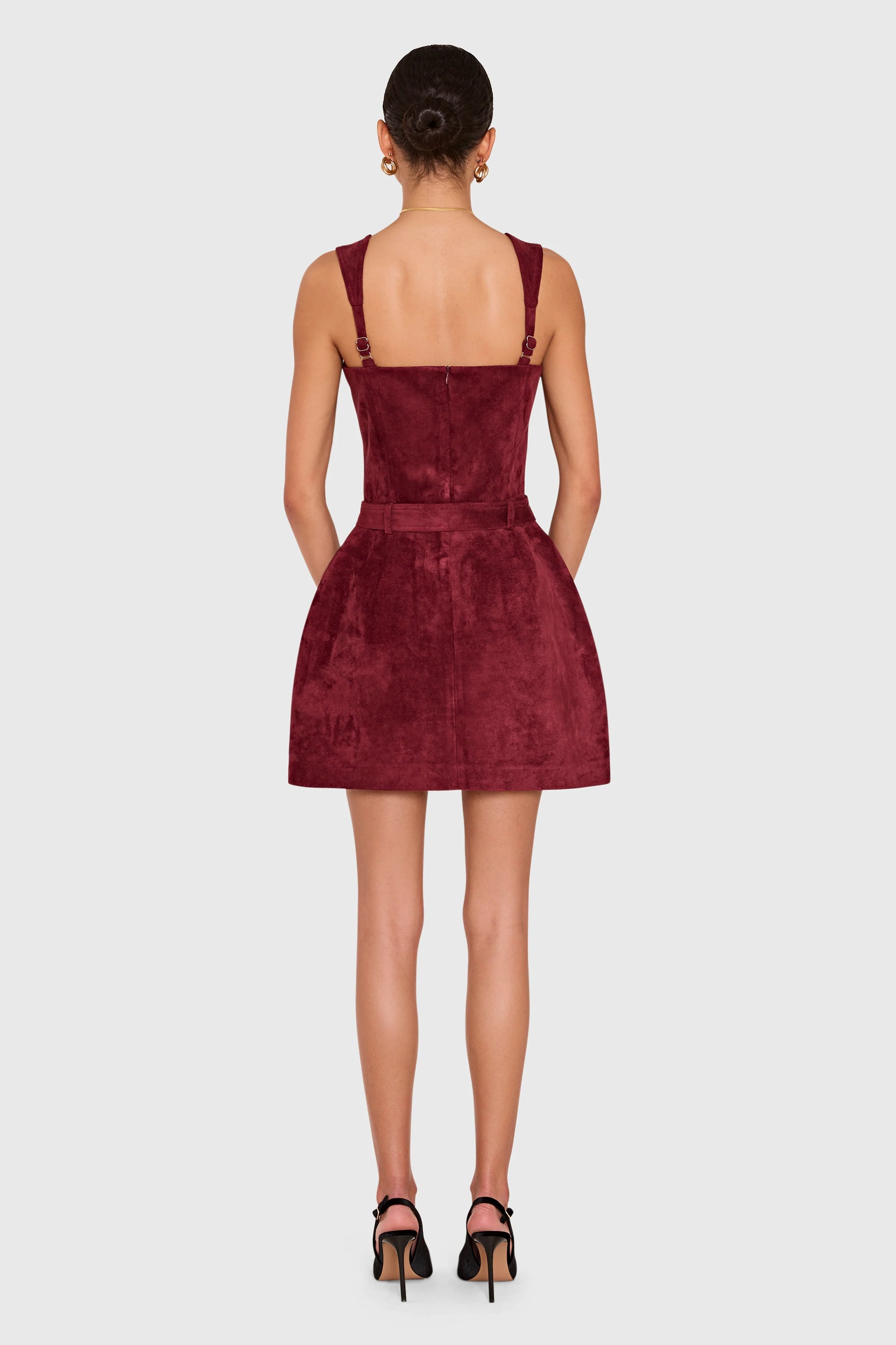 A woman viewed from the back wearing a sleeveless burgundy mini dress with shoulder straps and a fitted waist, paired with black high-heeled sandals, against a light gray studio background.
