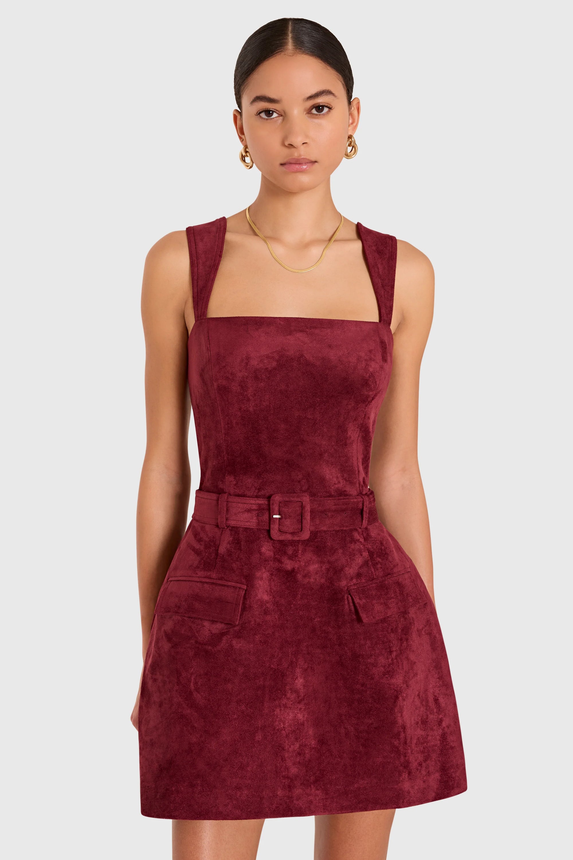 A model wearing a sleeveless, square-neck mini dress in a deep burgundy velvet-like fabric, featuring a matching belt with a square buckle and front patch pockets, set against a plain light gray background.