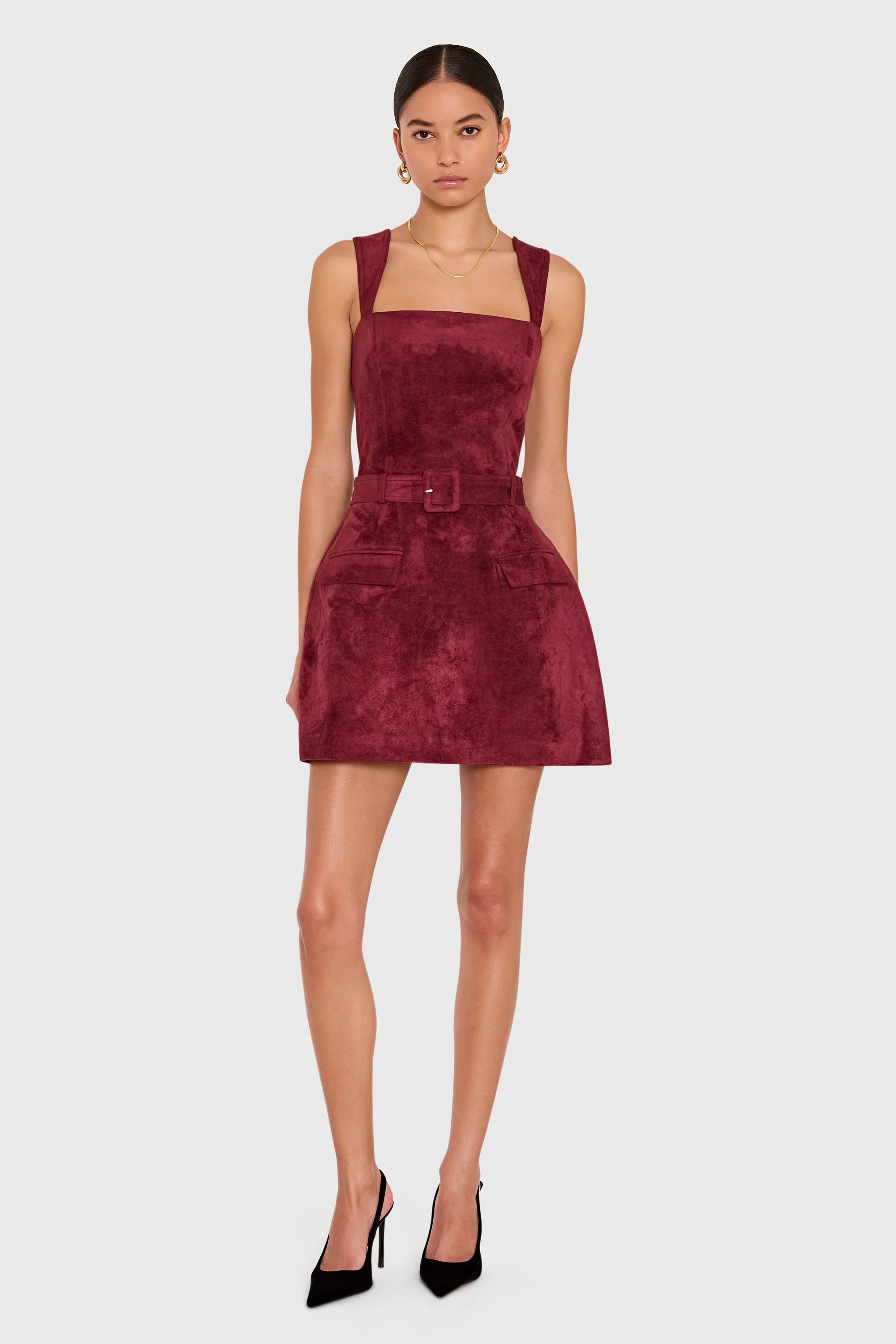 A model wearing a sleeveless, square-neck mini dress in a deep burgundy velvet fabric, styled with black high heels against a light gray studio background.