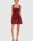 A model wearing a sleeveless, square-neck mini dress in a deep burgundy velvet fabric, styled with black high heels against a light gray studio background.