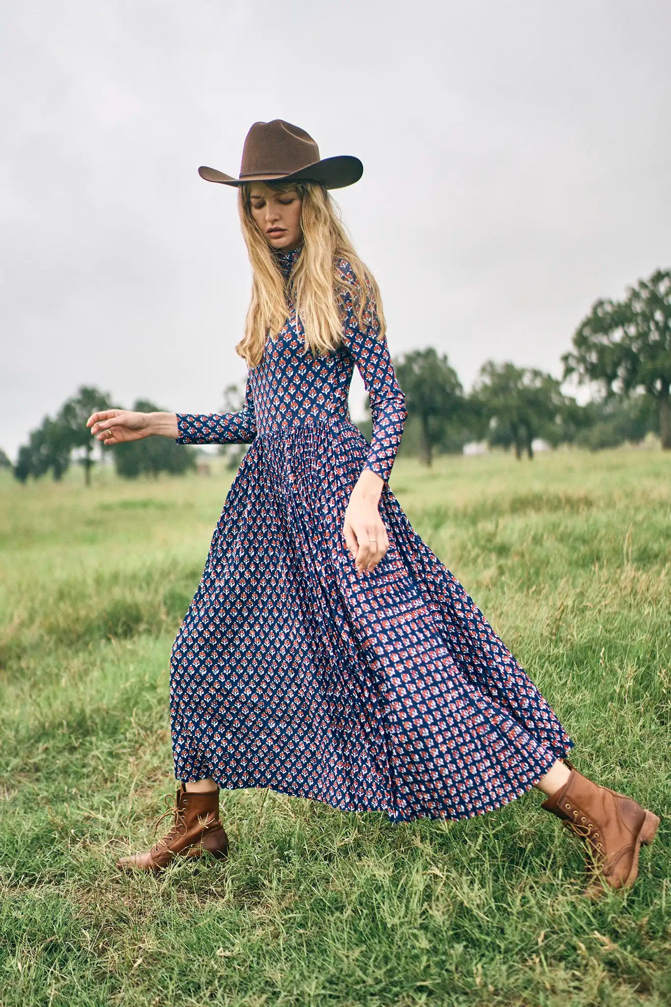 Woman in a long blue patterned dress and brown hat standing in a grassy field.