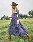 Woman in a long blue patterned dress and brown hat standing in a grassy field.