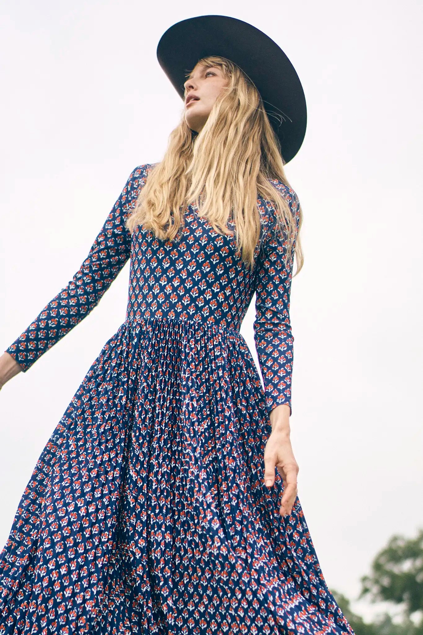 Woman wearing a patterned dress and black hat against a blurred natural background