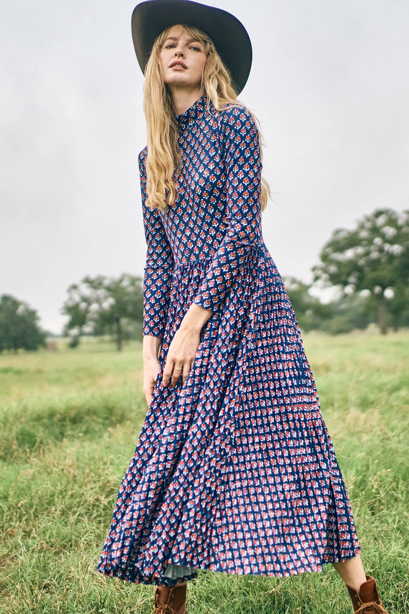 Woman in a patterned dress and wide-brimmed hat standing in a field.