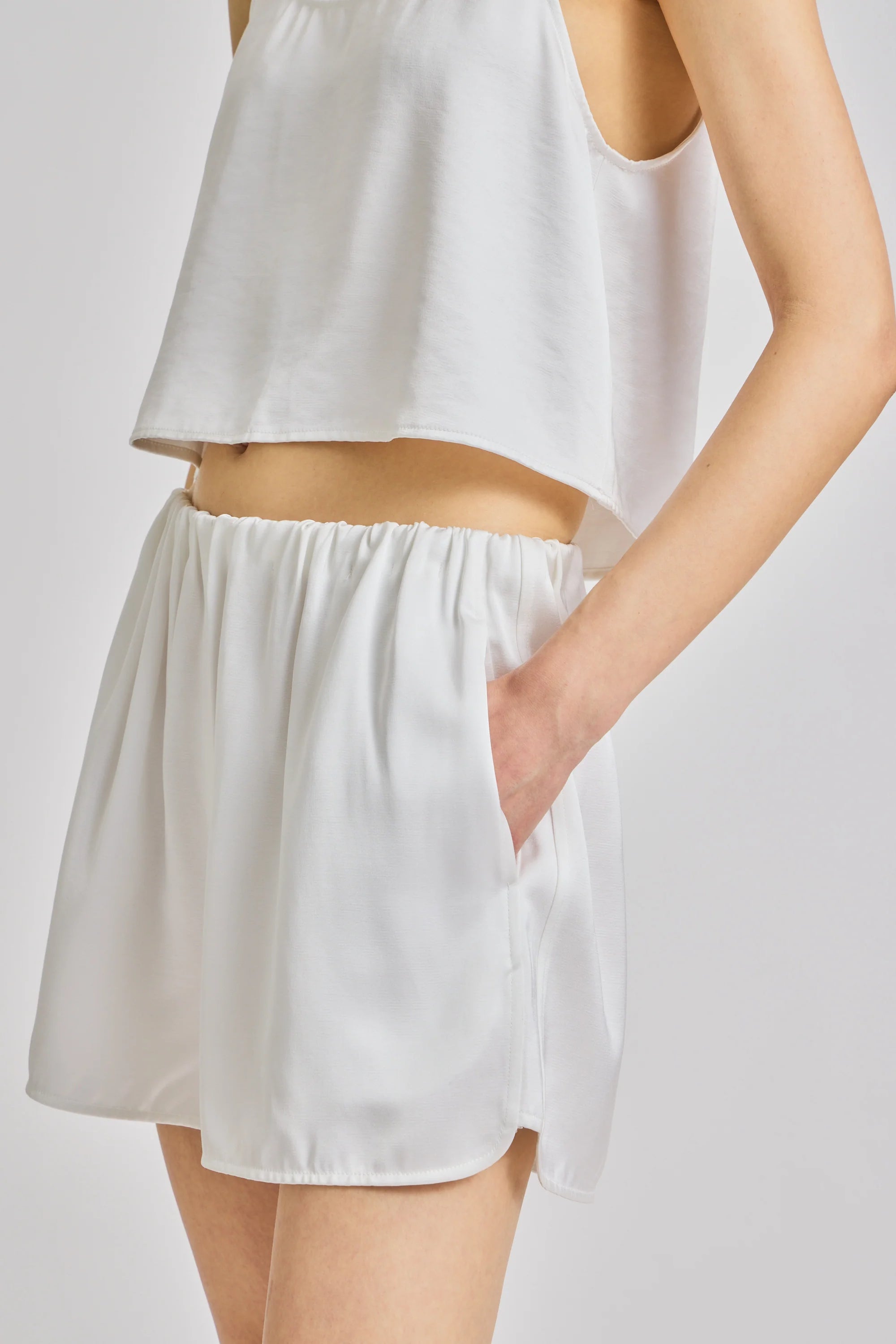 A close-up of a model wearing a white sleeveless crop top and matching white shorts with side pockets, set against a plain light gray studio background.