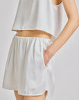 A close-up of a model wearing a white sleeveless crop top and matching white shorts with side pockets, set against a plain light gray studio background.