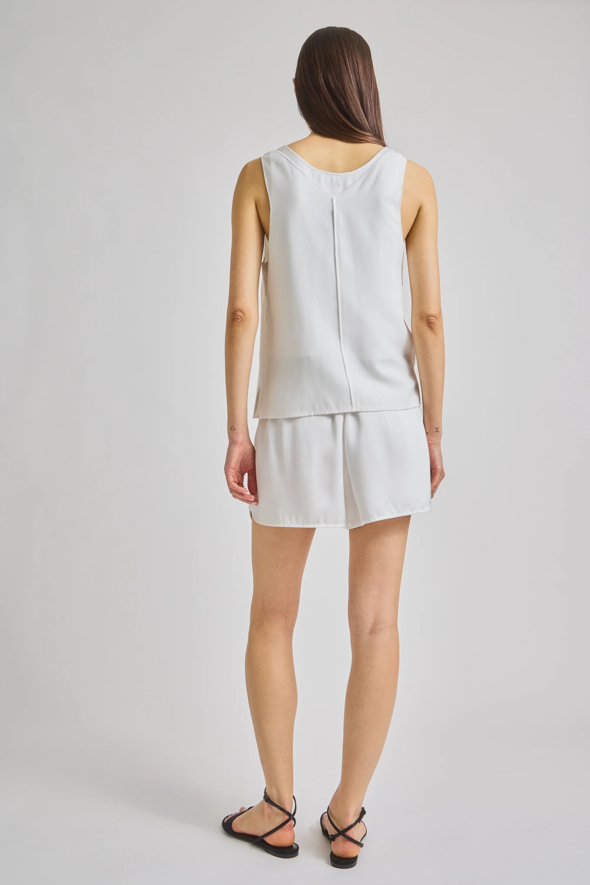 A woman shown from the back wearing a sleeveless white top and matching white shorts against a light gray studio background.