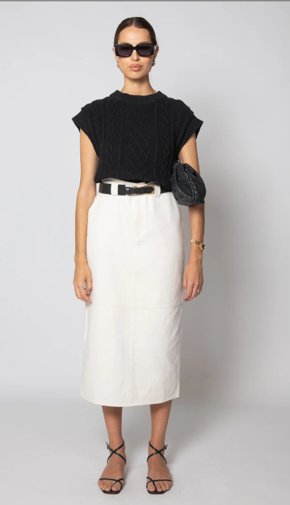 A model stands against a light gray studio background wearing a black short-sleeve textured knit top, a white midi skirt with a black belt, black sunglasses, black strappy heels, and a black woven crossbody bag.