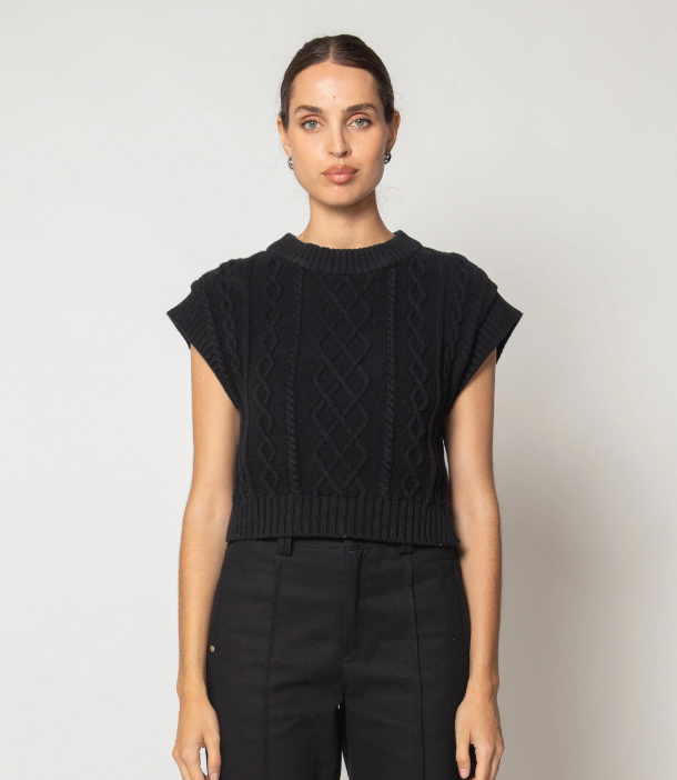 A woman modeling a black short-sleeve knit sweater with textured cable and diamond patterns, paired with black trousers, against a plain light gray background.