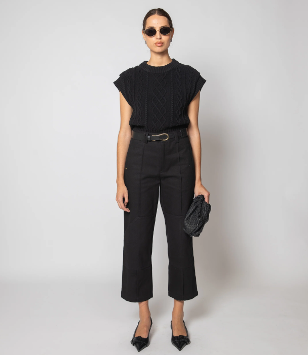 A model stands against a light gray studio background wearing a black short-sleeve textured knit top, black cropped trousers with a black belt, black pointed flats, sunglasses, and holding a black woven clutch.