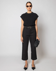 A model stands against a light gray studio background wearing a black short-sleeve textured knit top, black cropped trousers with a black belt, black pointed flats, sunglasses, and holding a black woven clutch.