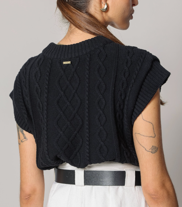 A woman shown from the back wearing a black cable-knit cropped sweater with short sleeves, paired with white pants and a black belt, against a plain light gray studio background.