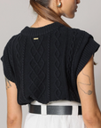 A woman shown from the back wearing a black cable-knit cropped sweater with short sleeves, paired with white pants and a black belt, against a plain light gray studio background.