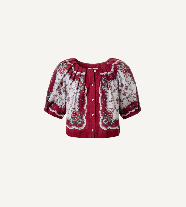 A short-sleeve button-front blouse with a deep V-neck, featuring a red base with a white floral and paisley pattern, displayed on a light cream background.