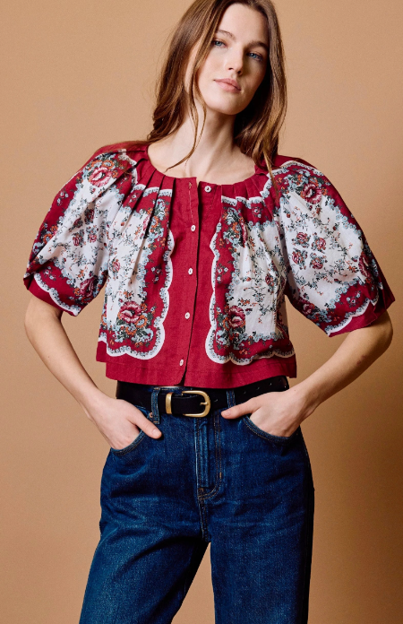 A woman modeling a short-sleeved blouse with a red and white floral pattern, paired with dark blue jeans and a black belt, set against a solid tan background.