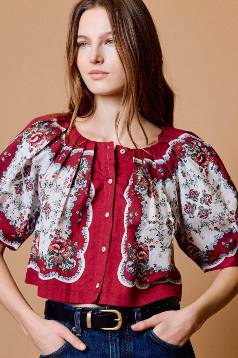 A woman wearing a red and white floral puff-sleeve blouse with a button front, paired with blue jeans and a black belt, against a solid tan background.
