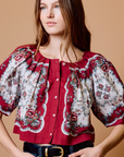 A woman wearing a red and white floral puff-sleeve blouse with a button front, paired with blue jeans and a black belt, against a solid tan background.