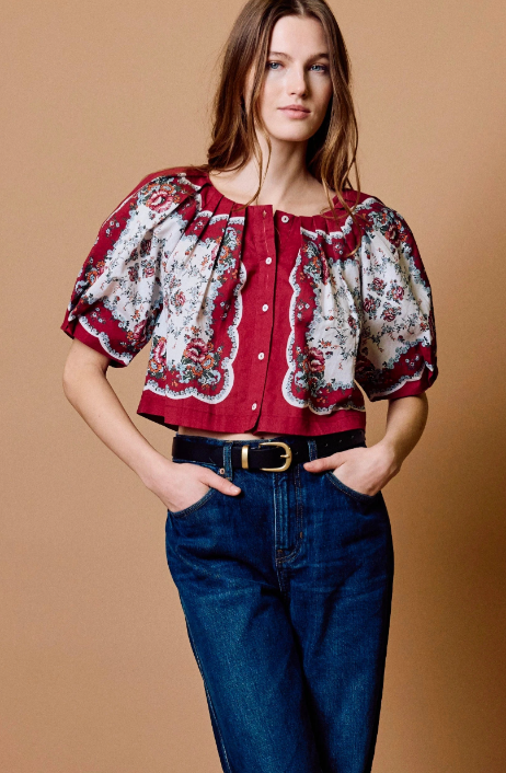 A woman modeling a short-sleeve blouse with a red and white floral pattern, paired with dark blue jeans and a black belt, against a solid tan background.