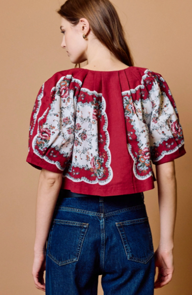 A woman shown from the back wearing a red cropped blouse with dramatic puff sleeves and a white floral print, paired with blue jeans, against a solid tan background.