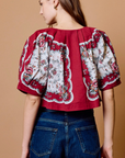 A woman shown from the back wearing a red cropped blouse with dramatic puff sleeves and a white floral print, paired with blue jeans, against a solid tan background.