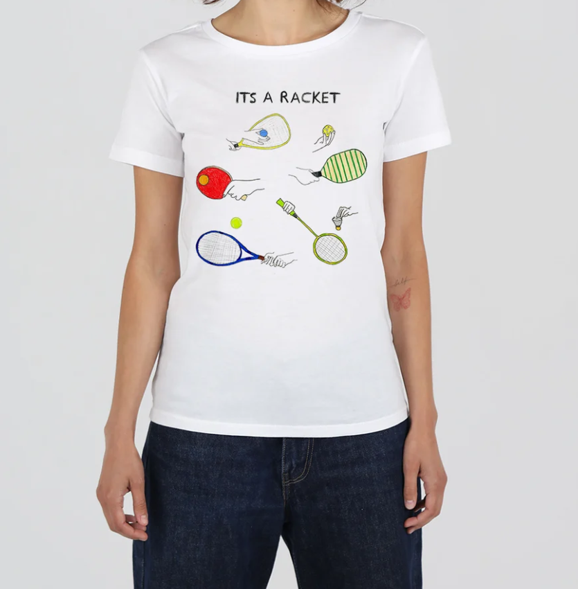 A woman wearing a white short-sleeve t-shirt with a colorful tennis-themed graphic that reads 'ITS A RACKET' above illustrated rackets and tennis balls, paired with dark blue jeans against a light gray background.