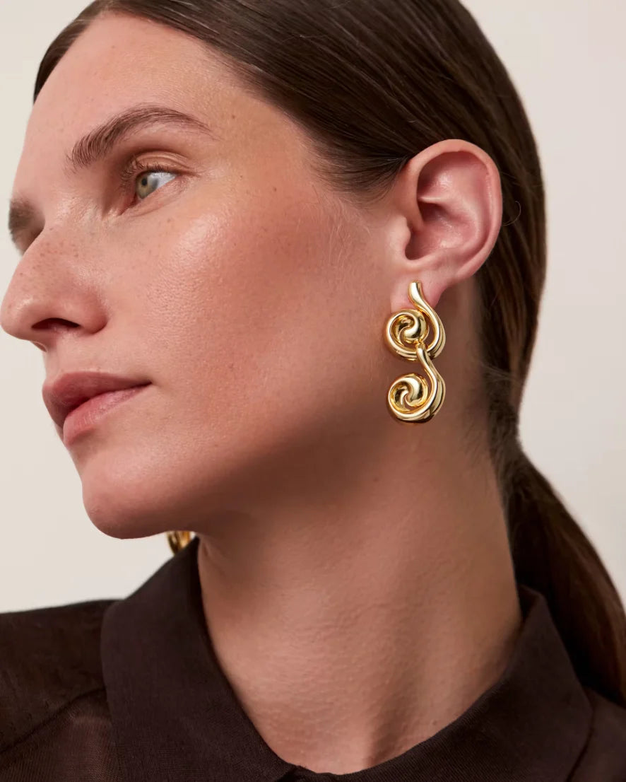 A close-up side profile of a woman wearing a gold spiral drop earring, showing the earring against her ear and neck.