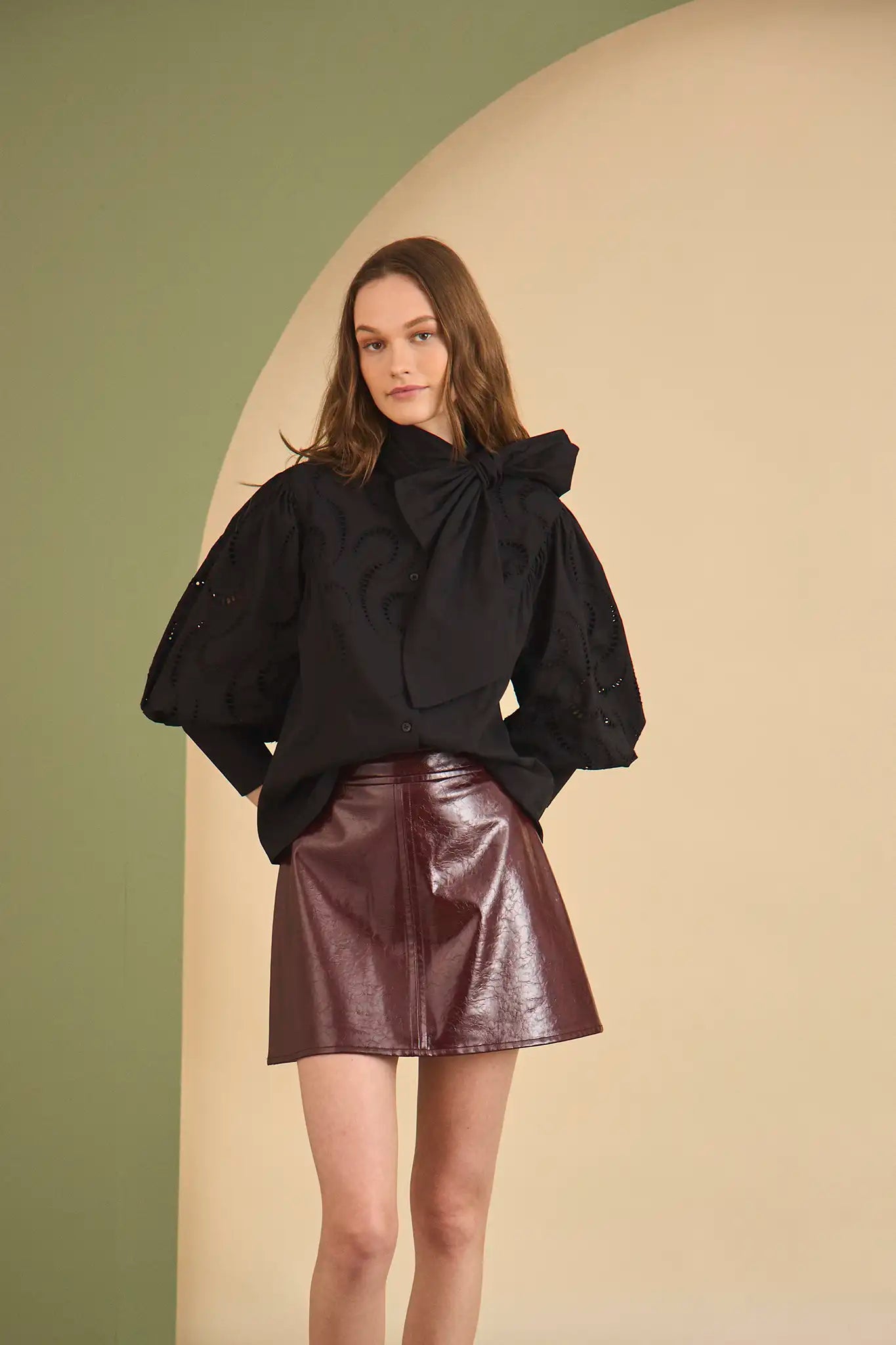 Woman wearing a black top with a bow and a shiny purple skirt against a beige and green background