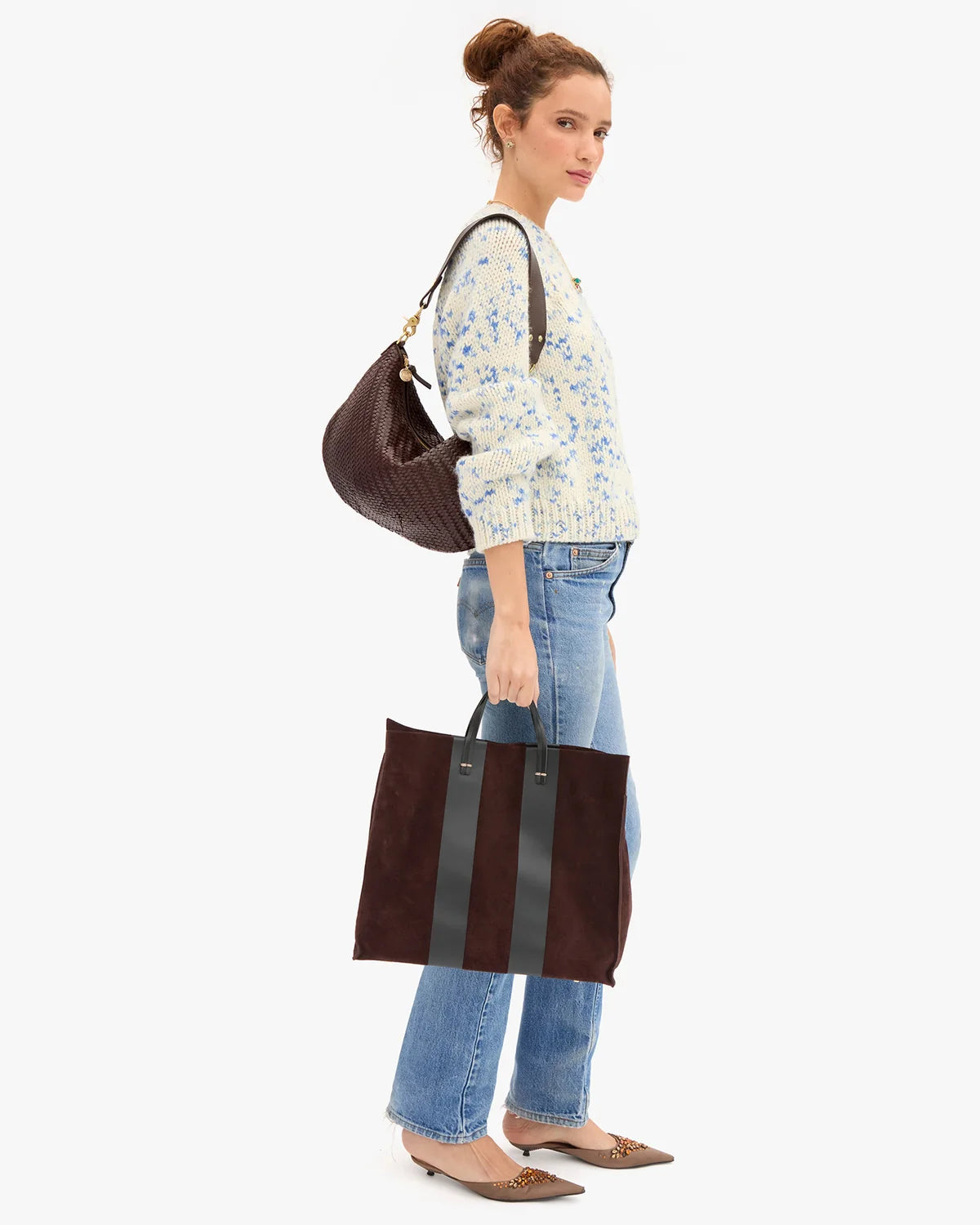 A woman stands in profile wearing a cream knit sweater with a blue floral print, blue jeans, and brown pointed flats, while carrying a dark brown woven shoulder bag and holding a dark brown tote bag with a vertical gray stripe. She is photographed against a clean, light gray studio background.