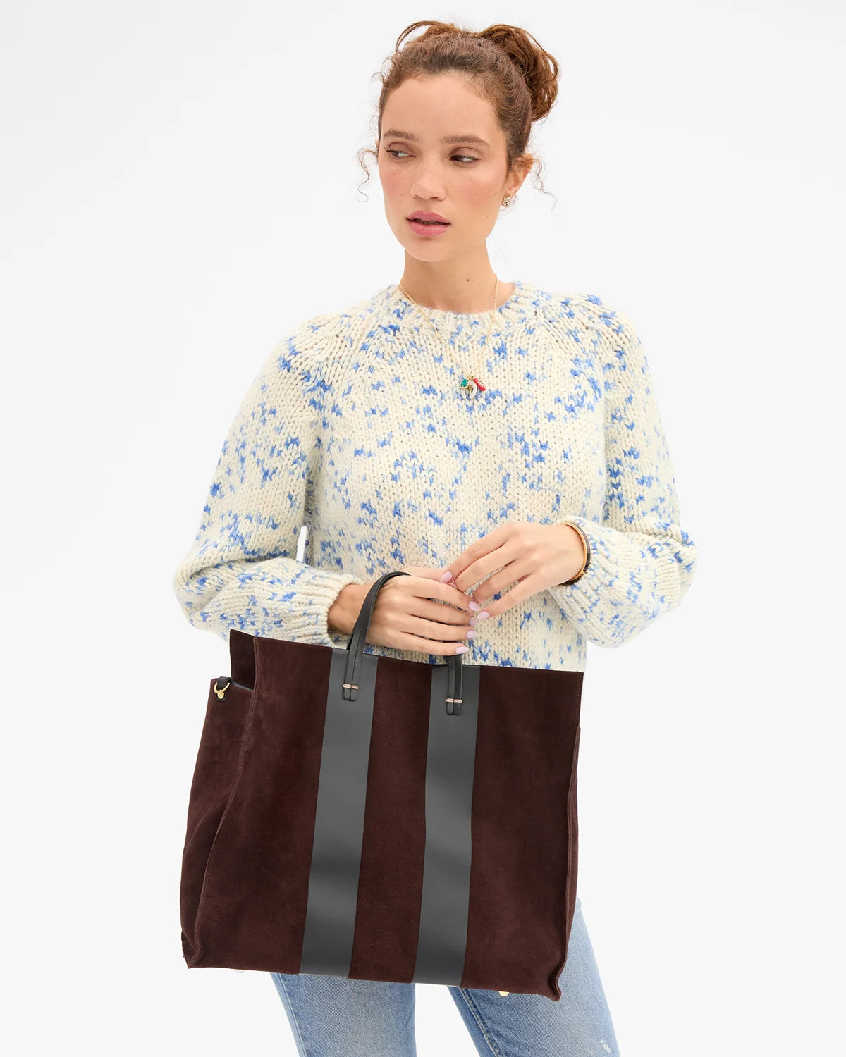 A woman wearing a cream knit sweater with a blue floral pattern and light blue jeans, holding a large dark brown suede tote bag with black leather handles and vertical black leather stripes against a plain white background.